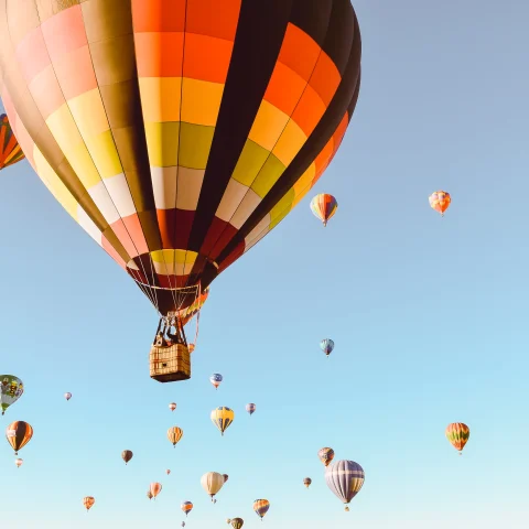 Red, yellow, white, orange, and black hot air balloon floats in the sky with other hot air balloons in the background in a clear blue sky