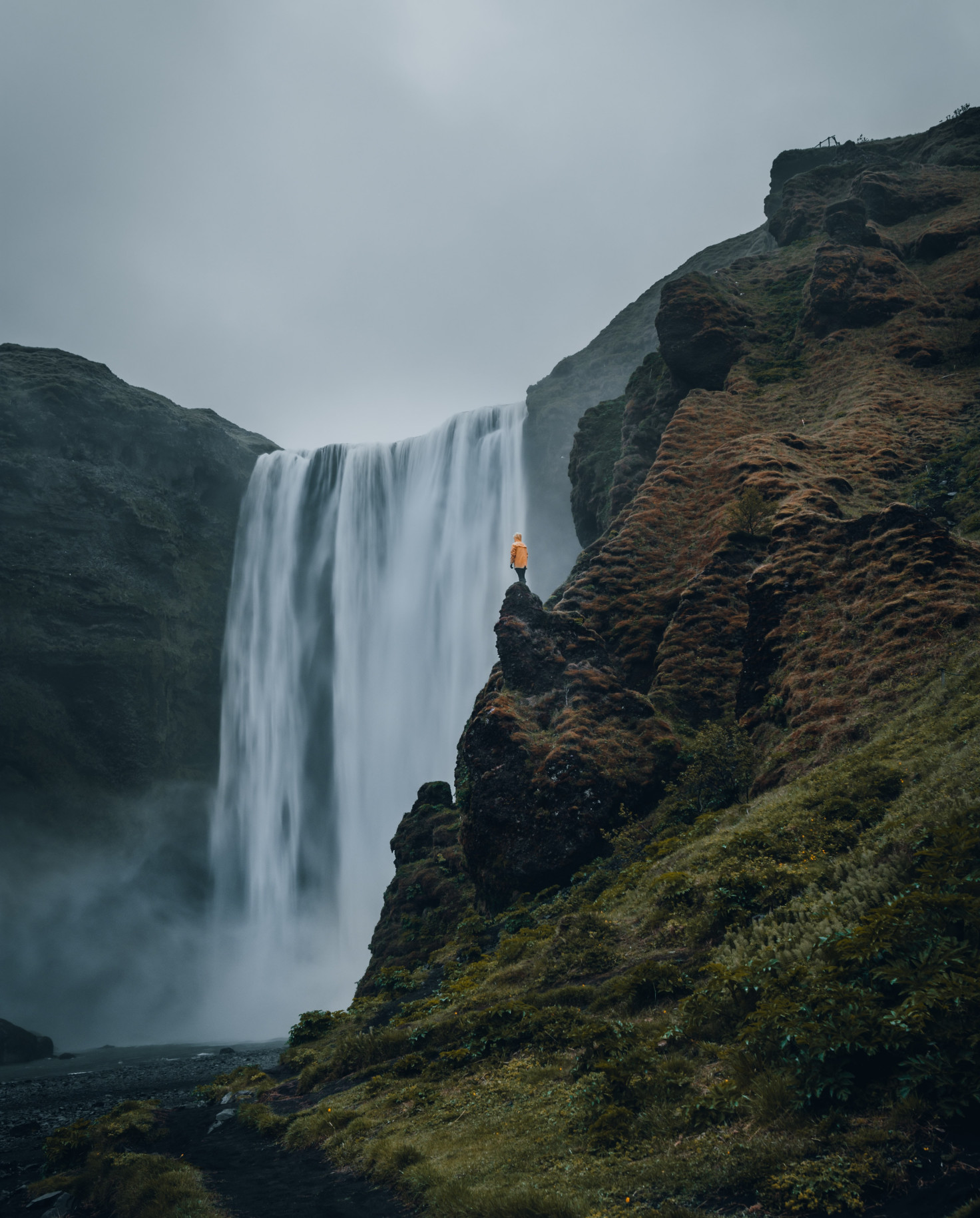 Person wearing orange jacket standing in front of waterfall on a cloudy day