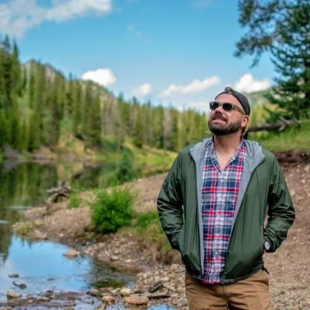 Travel Advisor Chris Pacal in a plaid shirt and green jacket in front of green trees and a lake.