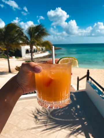 A hand presents a refreshing cocktail against a stunning beach backdrop, evoking a sense of tropical relaxation.