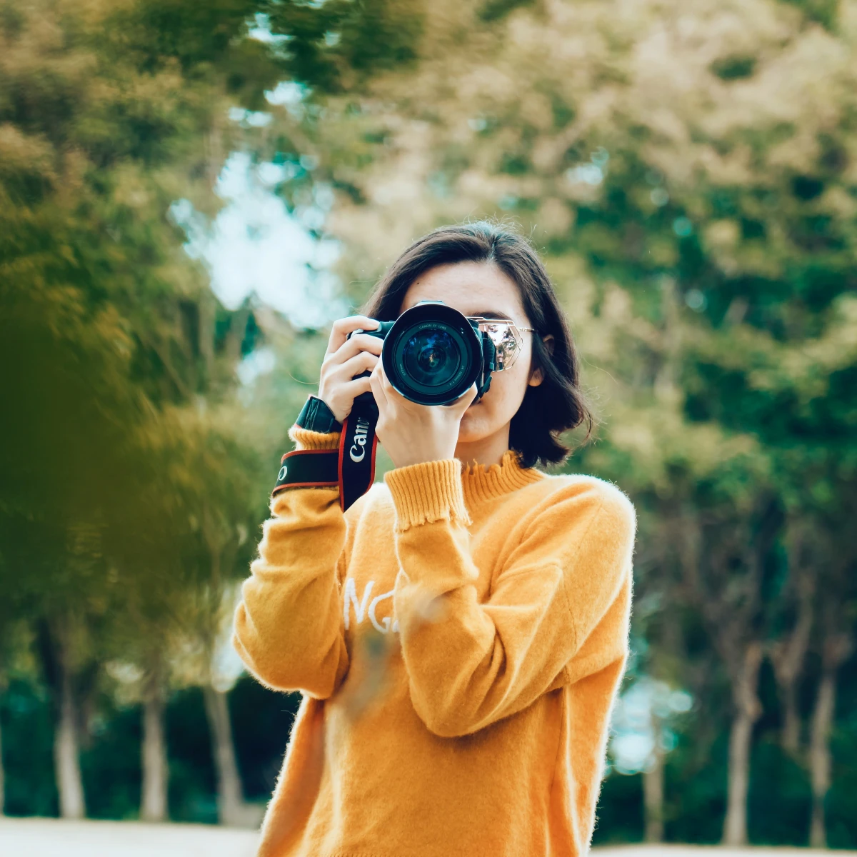 Woman wearing orange sweatshirt holding camera with trees in background