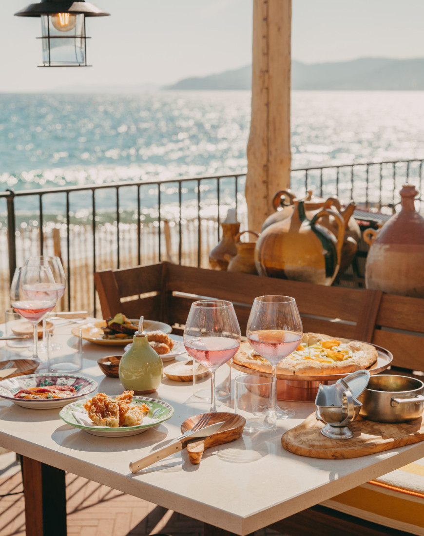 a seaside wooden table filled with wine glasses and colorful food