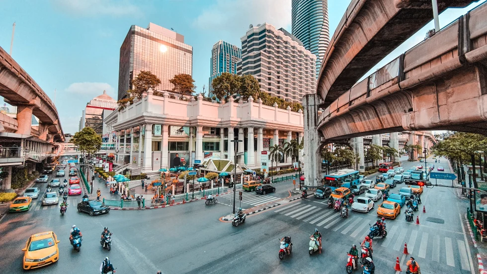 A street view of Bangkok, Thailand with yellow taxis, motorcycles, cars, and tan buildings, white crosswalks and concrete sky bridges.
