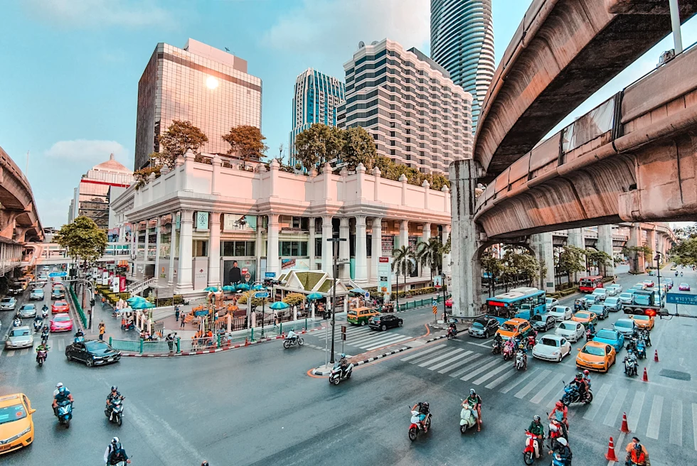 A street view of Bangkok, Thailand with yellow taxis, motorcycles, cars, and tan buildings, white crosswalks and concrete sky bridges.