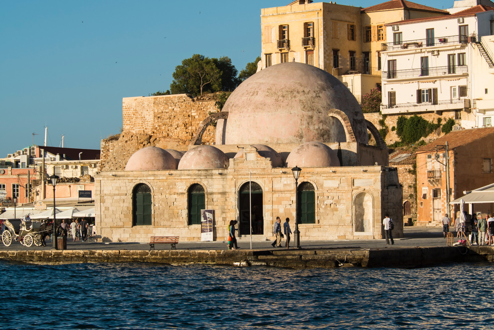 blue water and skies with white and tan ancient rounded buildings and a green leafy tree Crete old city Heraklion