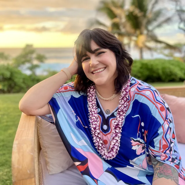 A woman sitting on a chair with a colorfully printed dress outside with palm trees and grass in the background. 