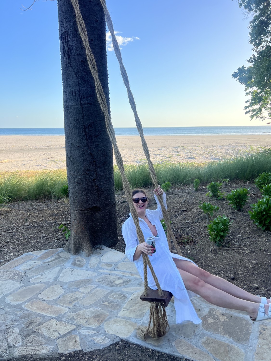 Travel advisor Emily in a white dress posing on a low swing with a long rope in front of a beach on a sunny day