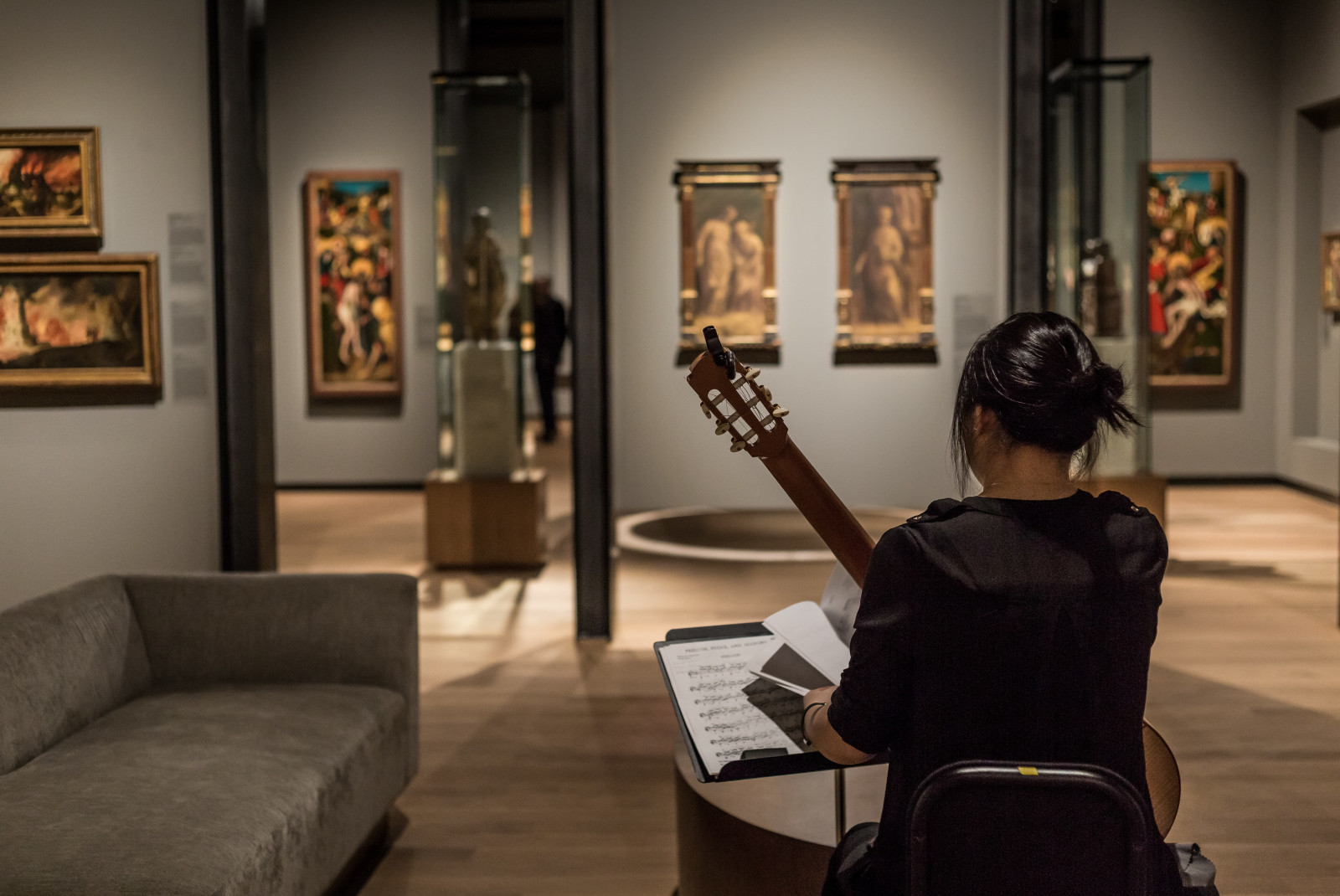 A woman from behind in black holding a guitar in a museum with six colorful painting and a tan couch
