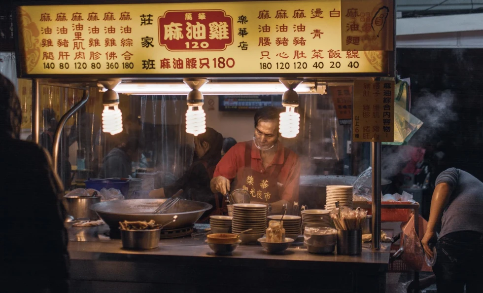 a man makes food in a food cart at night with steam rising from hot plates and a large wok