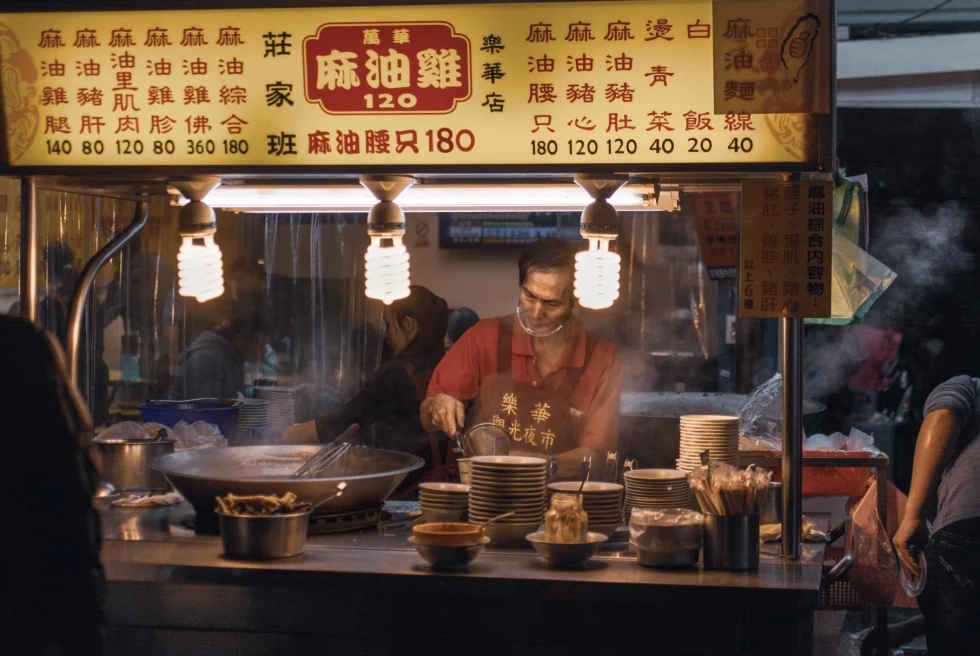 a man makes food in a food cart at night with steam rising from hot plates and a large wok