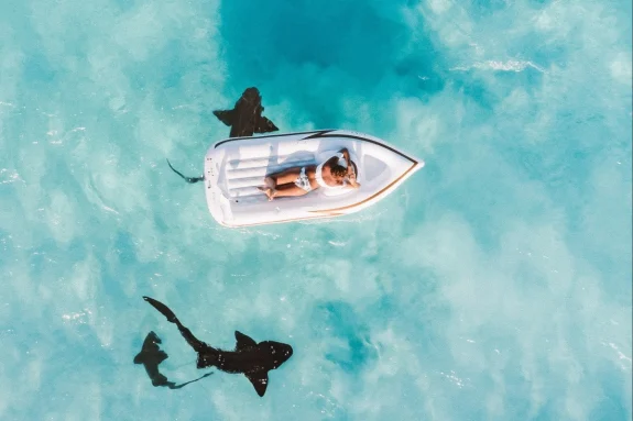 Girl lounging on a small boat floating idly in remote calm and clear waters as sharks swim underneath. 