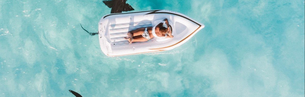 Girl lounging on a small boat floating idly in remote calm and clear waters as sharks swim underneath. 