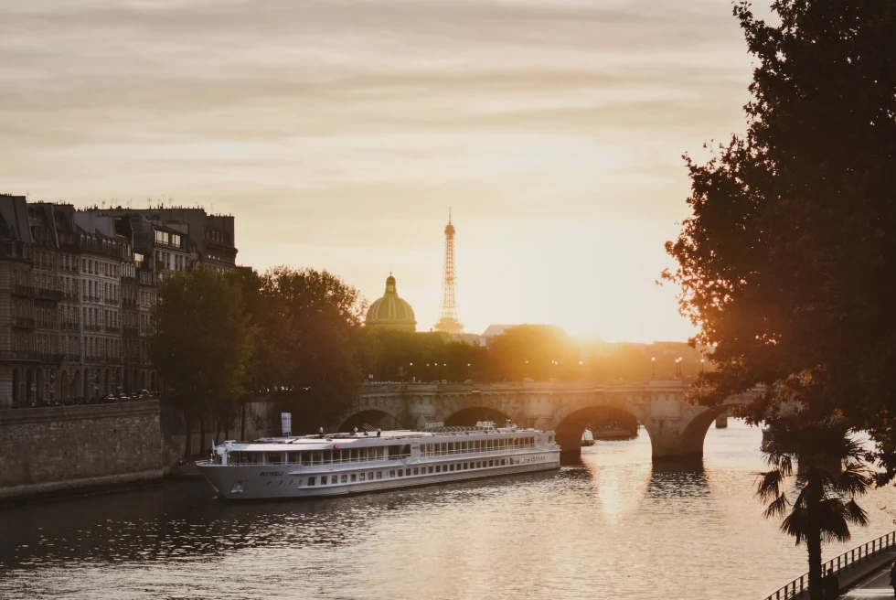 A reflective river with the golden sun setting and a large white boat and bridge in the distance with shadowed trees on the path along the water