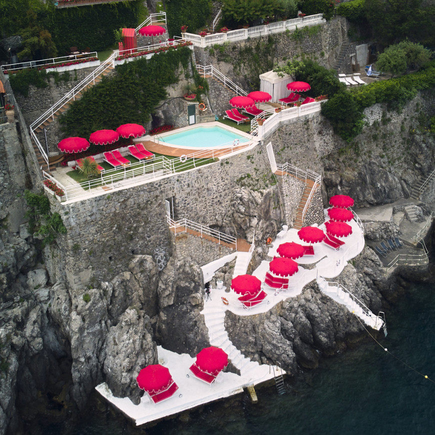 aerial view of a cliffside beachclub dotted with red umbrellas and red lounge chairs