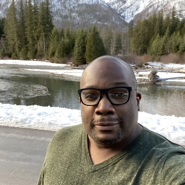 Travel Advisor Brian Hooper in a green sweater in front of green trees and snow capped mountains.