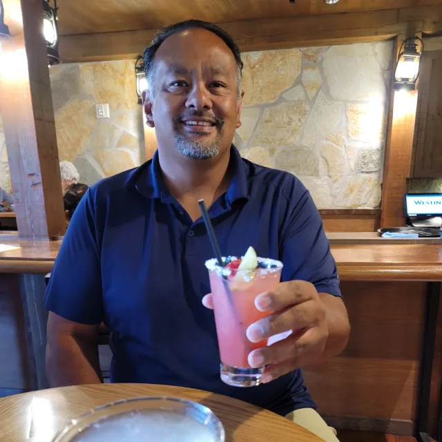 Travel Advisor John Chamberlain holds up a pink cocktail at a bar with a map all the wall behind him