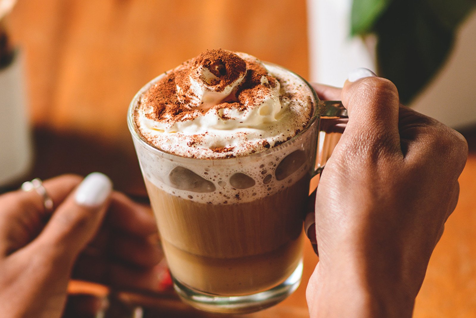 Hot chocolate in a glass mug with whipped cream and cinnamon held by a woman's hands with white nail polish and a wooden floor and green plant in the background