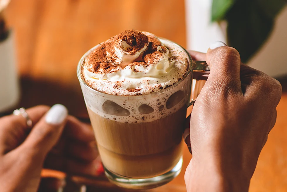 Hot chocolate in a glass mug with whipped cream and cinnamon held by a woman's hands with white nail polish on cold winter day.