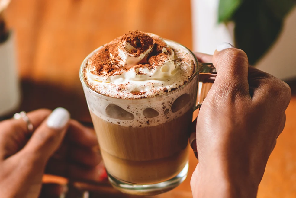 Hot chocolate in a glass mug with whipped cream and cinnamon held by a woman's hands with white nail polish on cold winter day.