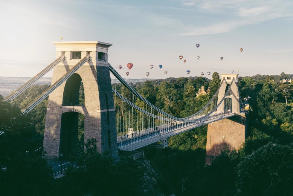 hot air balloons over suspension bridge surrounded trees 