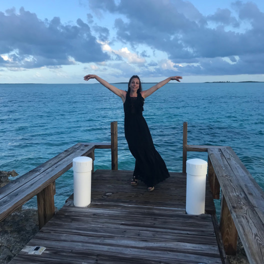 Woman standing in black dress on beach dock with her arms in the air and smiling