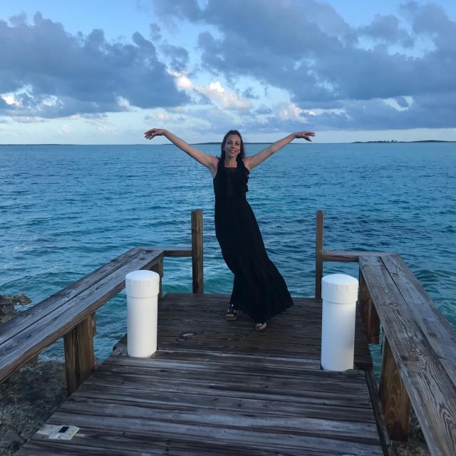 Woman standing in black dress on beach dock with her arms in the air and smiling