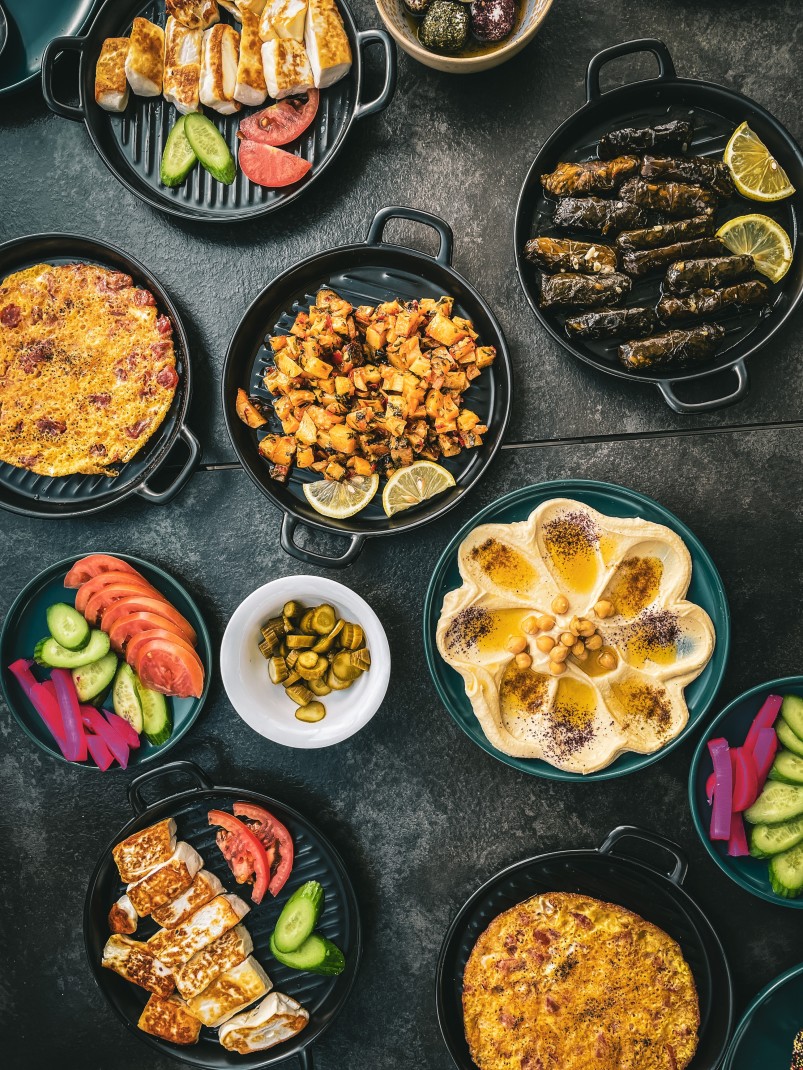 A spread of Middle Eastern food in Jordan, such as tomatoes, cucumbers, pickled onions, and hummus, dolmas with yellow lemon and tan chicken and rice served in black cast iron skillets.