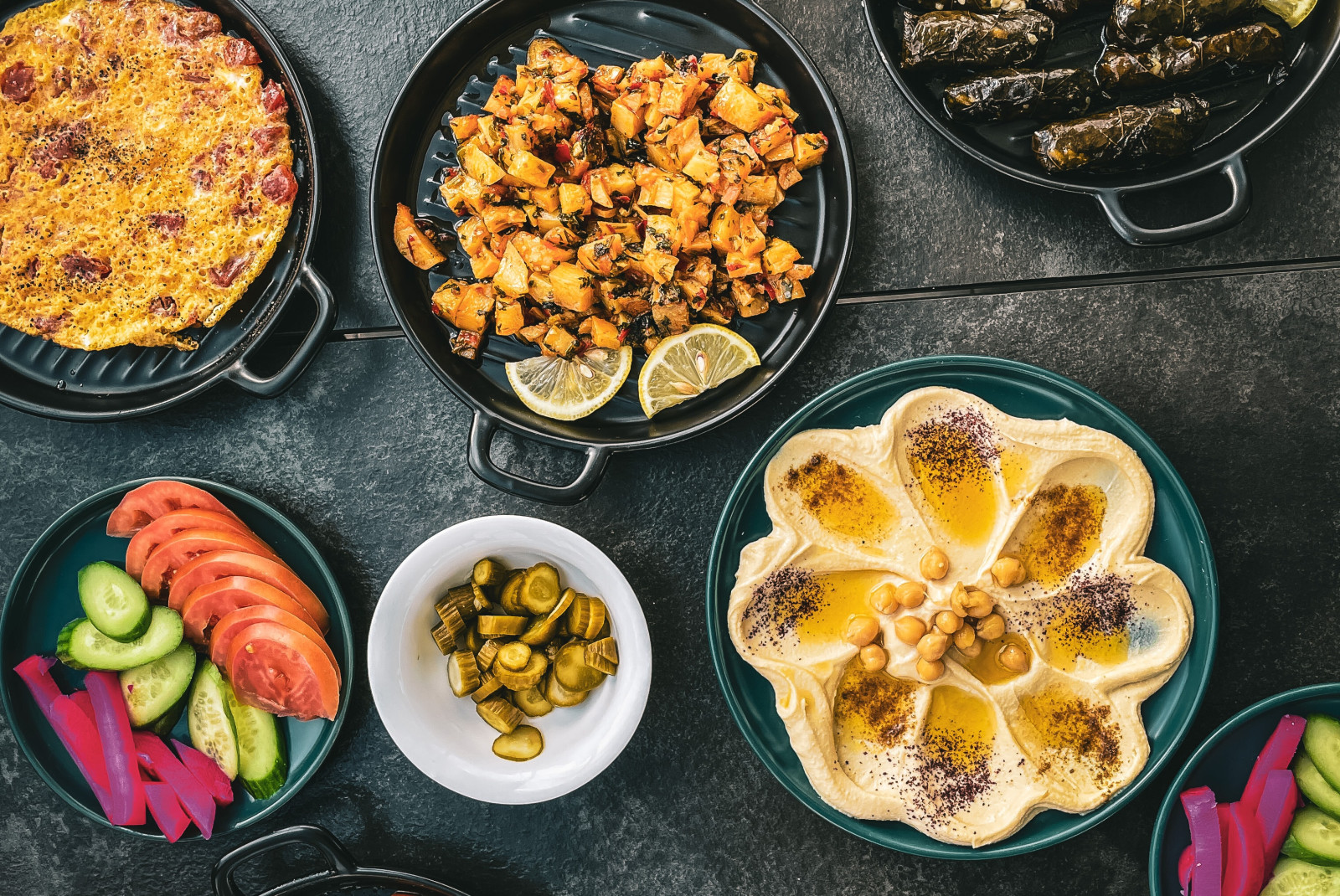 A spread of Middle Eastern food in Jordan, such as tomatoes, cucumbers, pickled onions, and hummus, dolmas with yellow lemon and tan chicken and rice served in black cast iron skillets.