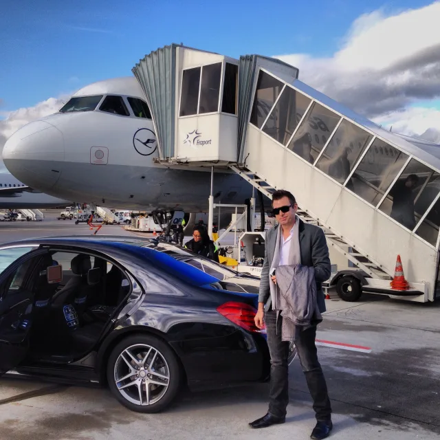 Anthony Losanno in a suit posing near a car and airplane on a runway.