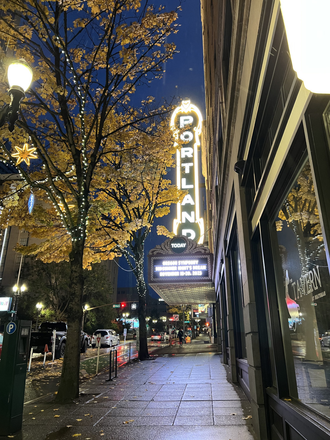 A view of a theatre sign that says Portland it lit up gold lettering next to a city sidewalk and trees. 
