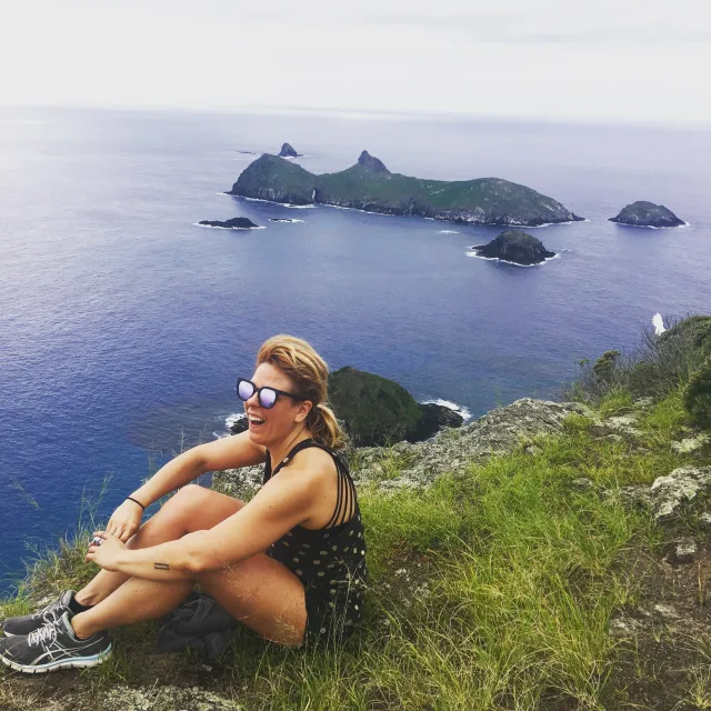 travel advisor Adrienne Dwulet sits on the top of a grassy cliff over the ocean wearing sneakers and reflective sunglasses