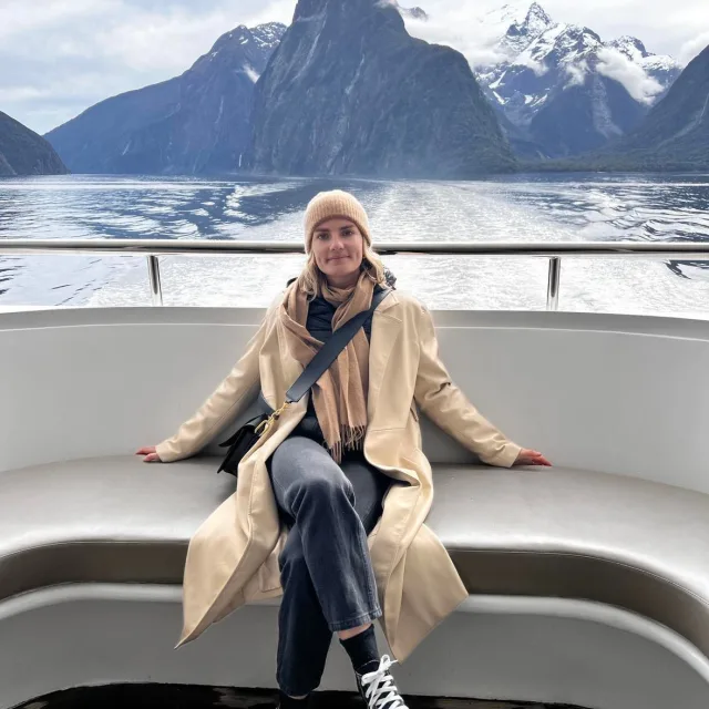woman wearing tan coat sitting on a boat with mountains in the background