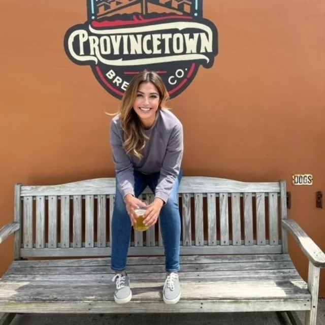 Vannesa Tiemann sitting on a bench in front of an orange wall with a sign that says 'Provincetown'.