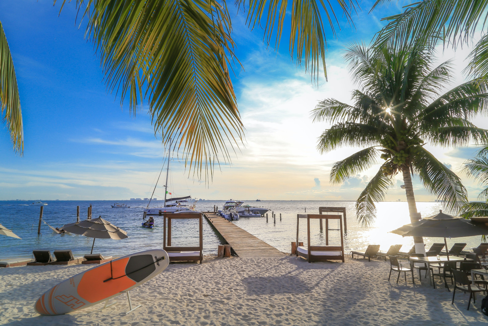 Pier leading from beach into ocean with boats in the water and palm tree in the foreground