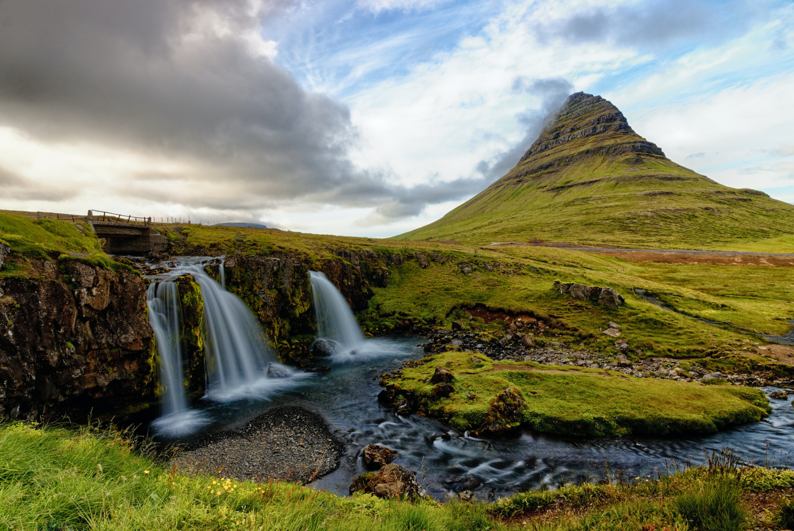 Two waterfalls next to large green mountain with clouds in the sky during daytime