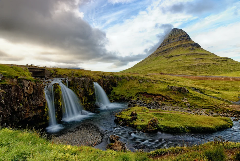 Two waterfalls next to large green mountain with clouds in the sky during daytime