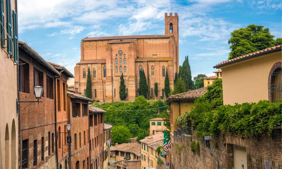 A tan-orange church surrounded by brown buildings and green trees in Siena, Italy.