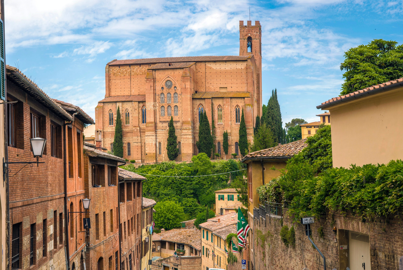 A tan-orange church surrounded by brown buildings and green trees in Siena, Italy.