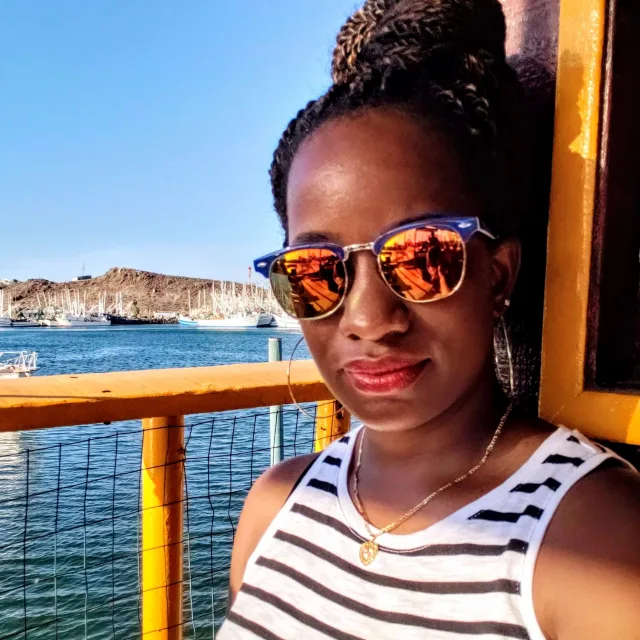 Travel Advisor Chyla Dalton-Nava in a white shirt with black stripes, sunglasses with sea in the background. 