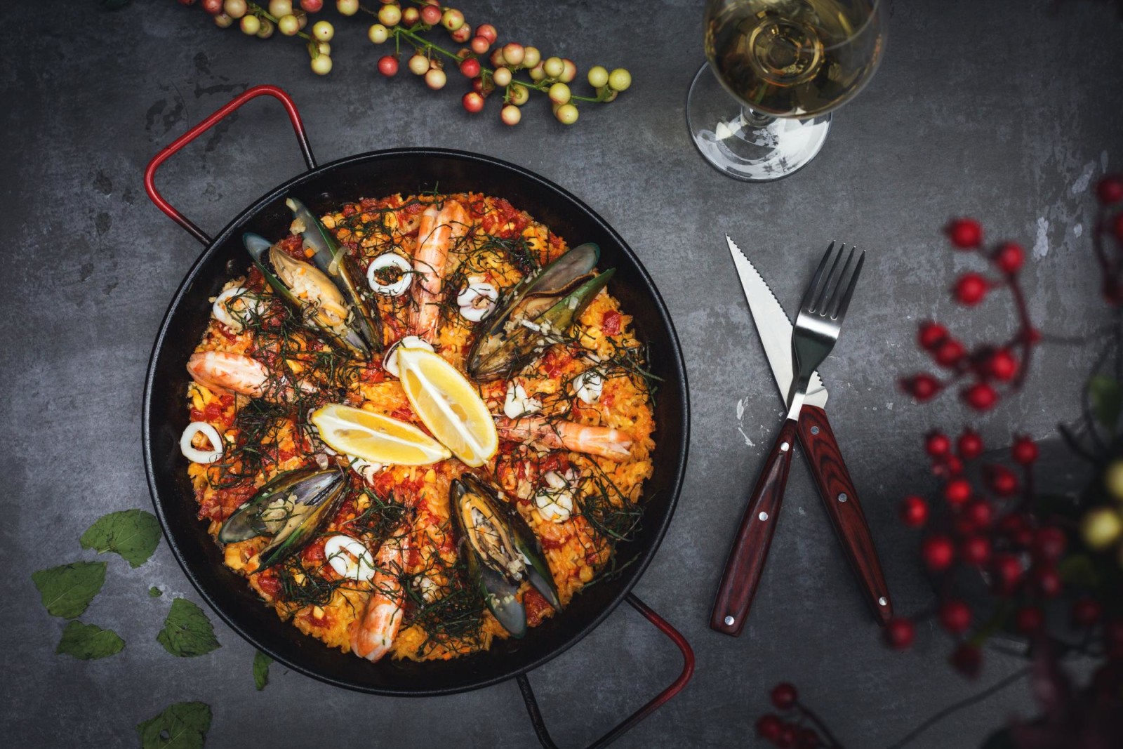Hot paella with mussels, shrimp and squid in pan with utensils and drink on the side on charcoal table.