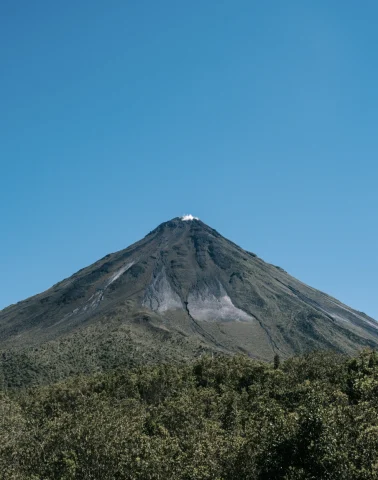 A large volcano surrounded by trees and blue sky. 