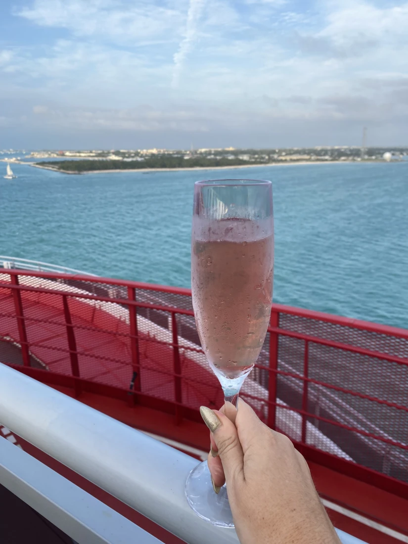 A picture of a hand holding a champagne flute with a light pink liquid inside of it. There is a red railing and view of the blue sea in the background. 