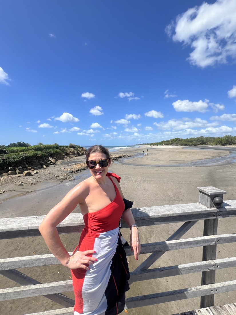 Travel advisor Emily standing on a wooden deck in a red, white and black dress in front of a beautiful beach