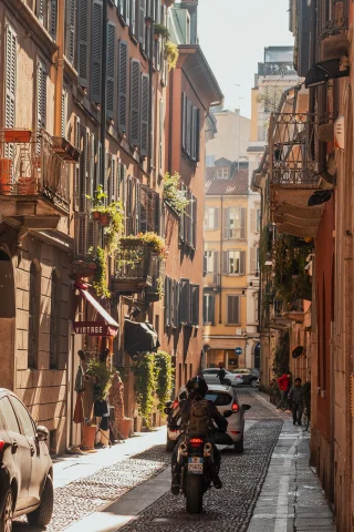 A cobblestone street in Milan with a scooter and some cars. 