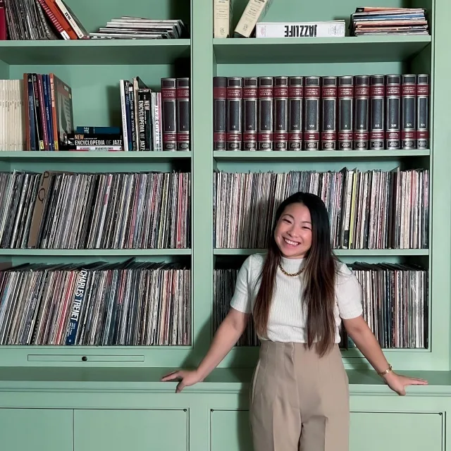 Travel Advisor Megan Soh poses in front of a light green book shelf wearing khaki trousers and a white sweater