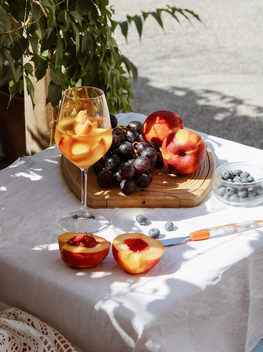 a glass of white wine with a plate of grapes and peaches resting on a white tablecloth