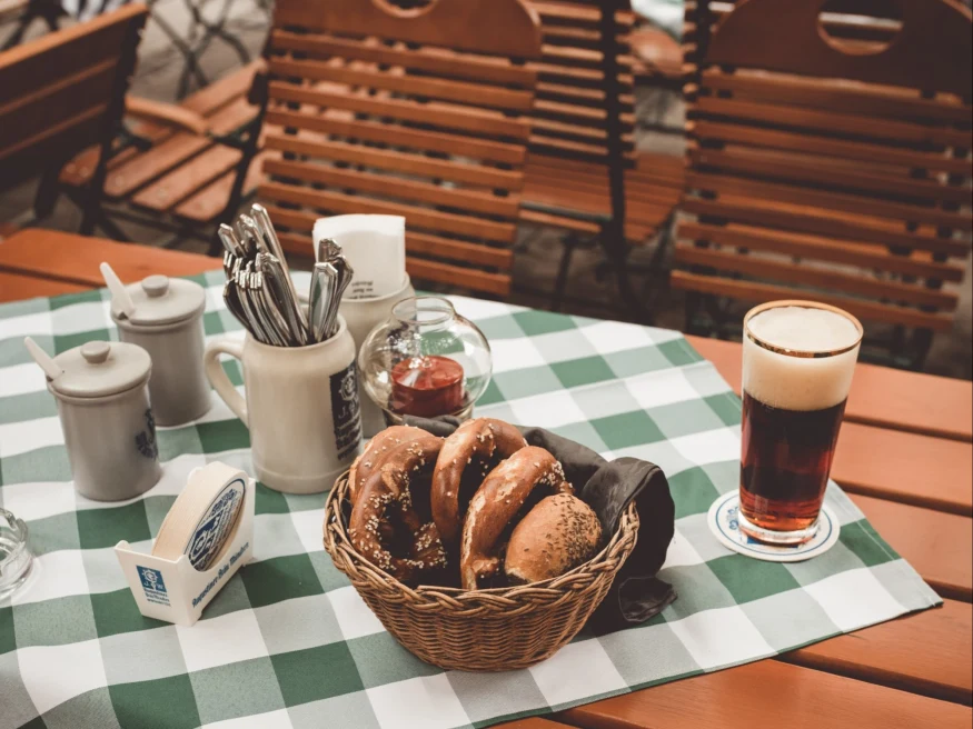 Pastries in basket with beer and silverware on plaid table cloth on picnic table.