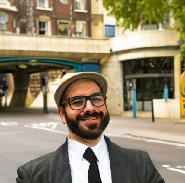 Phelipe wearing glasses, a suit and a hat while smiling and standing outside in front of a city street. 