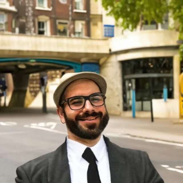 Phelipe wearing glasses, a suit and a hat while smiling and standing outside in front of a city street. 