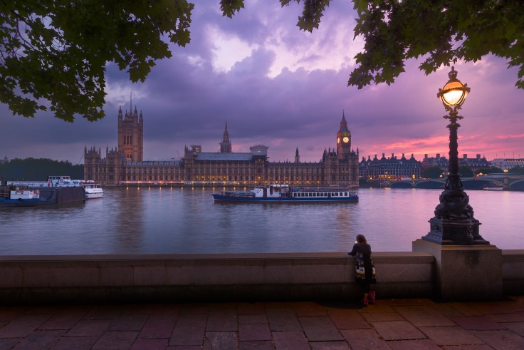 Parliament in London, England at sunset with pink and purple dark clouds and a woman standing across the river next to a lamp post.